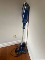 Full vacuum cleaner standing upright showing overall design, color, and included charger cord on floor.