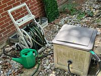 Two white plastic hose reels with green garden hoses, a green watering can, and a white plastic storage box with hose attached sitting outdoors on rocks near a brick wall.