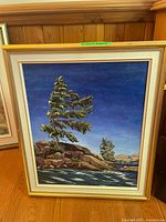 Full view of framed acrylic painting featuring a pine tree, rocks and water under deep blue sky.