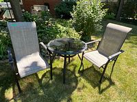 Two canvas and aluminum patio chairs and a round glass-topped outdoor table set on grass, showing wear and use.