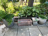 Four outdoor planters lined up on a patio with green leafy plants, a rectangular reddish-brown planter with ornate detail, and a large hanging fountain with visible damage in the background.