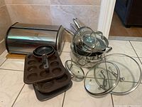 Wide view of lot with stacked Paderno pots with lids, metal bread box, wood cutting board, muffin pans, and two frying pans.