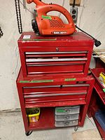 Front view of smaller and larger red tool chests stacked with a Black & Decker blower on top, showing drawer handles, wheels, and surface wear.