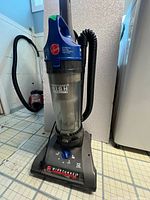 Front view of Hoover WindTunnel Techtronic upright vacuum cleaner showing blue handle, clear dust canister, power dial on base, and hose attachment.