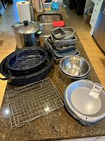 Countertop with various baking and roasting pans stacked, including a large black speckled roasting pan with wire rack, several small loaf and round pans, a large stainless steel pot with lid, and wire cooling racks.