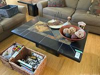 Photo of a square coffee table with tiled surface featuring black and brown geometric tile pattern, placed on a wooden floor between chairs and couches, holding decorative bowls and items.