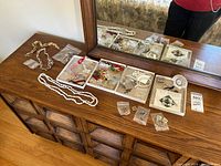 Full view of costume jewellery arranged on wooden dresser with mirror reflection. Includes pearl bead necklaces, boxed brooches, earrings, pins, and small clock.