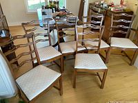 Six wooden dining chairs with light upholstered seats, arranged on a hardwood floor near a dining table and other furniture. Five side chairs and one captain's chair with arms visible.