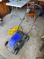 Blue and black Kobalt electric lawnmower on garage floor with yellow extension cord beside it, showing handle and wheels.