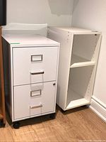 Photo shows one white metal filing cabinet with two drawers and one white wooden shelf unit side by side on a wooden floor against a white wall.