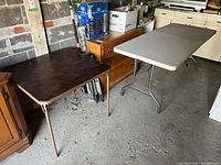 View of both folding tables side by side in a garage or storage area, showing brown vinyl square table and gray plastic rectangular table.