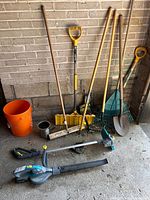 Photo of all gardening tools against a brick wall, includes blower, trimmer, rakes, shovel, bucket and broom.