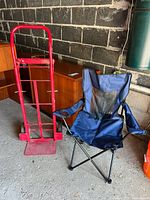 Red metal hand truck and blue folding picnic chair side by side in garage setting.