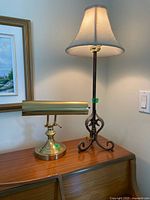 Wide shot showing both lamps side by side on a wooden surface with a painting in the background. Brass desk lamp with horizontal shade on left, tall metal lamp with fabric shade on right.