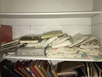 Shelf containing multiple stacks of folded linens including white, floral, and plastic-wrapped items.
