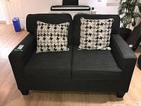 Front view of the dark gray upholstered loveseat with two patterned cushions on a light wood floor near a keyboard and potted plant.