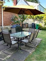 Full view of patio furniture set on stone patio, showing rectangular glass table, six sling patio chairs, and beige umbrella open over table