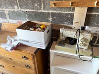 Sears Kenmore sewing machine placed on a white surface next to a white cardboard box filled with assorted sewing supplies including spools of thread and plastic bags containing smaller items.