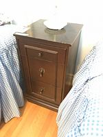 Three drawer wooden side table shown from slight left angle, featuring dark brown wood finish, one small top drawer with a knob, two bottom drawers with decorative keyhole handles.