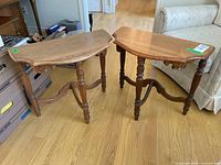 Two half moon style wooden side tables with beveled edges, turned legs, and carved aprons placed on wood flooring near sofa and boxes.