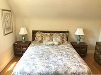 Pair of wooden side tables placed on each side of a bed with bedding and pillows. The tables have a dark brown wood finish and two shelves each. The tables appear lightly used and the bedding is not included in the lot.