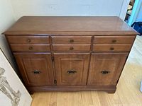 Front view of wood veneer sideboard showing three drawers and three doors with metal pulls, placed on wooden floor near a wall with adjacent framed artwork.