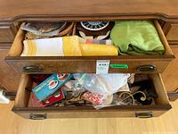 Two open drawers showing linens and assorted kitchen and serving items inside. Top drawer has mixed kitchen and table items including trivets, utensils, and cloths.