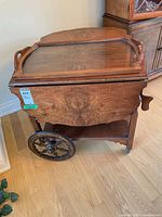 Side view of vintage wooden tea trolley showing drop leaf sides, decorative wooden wheels and lower glass tray