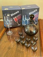 Photo showing the twelve Bohemia Diana smoked glass flutes in original boxes along with nine small smoked glass WMF Kristall liqueur glasses arranged on a wooden surface.