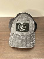 Front view of Klein Tools grey camo trucker hat showing logo patch, brim, and mesh back