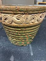 Close-up front view showing intricate carved swirl pattern adjacent to rim and green horizontal ridges below on the body of the pottery piece, highlighting the textured surface and color.