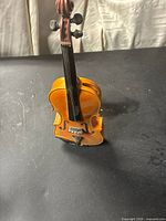 Front view of brown ceramic miniature violin figurine showing body, strings, tuning pegs, and bridge