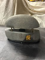Gray stone table lamp with rounded top placed on matching base showing gold leaf emblem on the front.