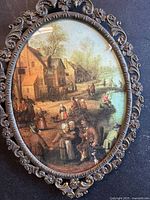 Full view of the oval vintage print depicting a Flemish village scene with peasants, horse-drawn carts, and buildings framed in an ornate bronze frame with floral and scrollwork details.