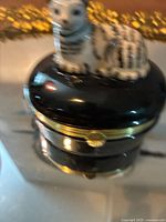 Close-up photo of the Limoges box showcasing the black glossy base, gold-tone clasp, and detailed cat figurine on top with black and white markings and green accent at neck.