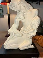 Front and slight side angle view of the cream ceramic sculpture showing the mother figure with two children on a black base, placed on a table with background items.
