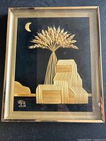 Full view of the framed straw art showing wheat sheaf, straw blocks, and crescent moon on black background in gold-tone metal frame.