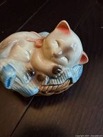 Angle view of the vintage ceramic cat figurine showing the sleeping cat curled on a textured pillow with soft blue and beige colors.