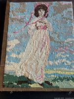 Full view of the vintage needlepoint embroidery with a woman in pink bonnet and dress against a cloudy blue sky and green grass background.