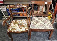 Two vintage wooden armchairs, one with a carved sculptural floral back, the other with a carved stylized X back, both with floral upholstered seats.