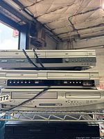 Stacked combo DVD and VHS players on metal rack, showing all three units from side angle.