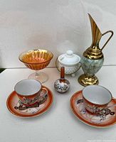 Photo showing two Asian tea cups and saucers, carnival glass compote, milk glass sugar dish, small ceramic vase, and brass accented vase