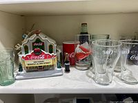 Wide view of Coca-Cola collectibles including light-up Christmas theater, multiple Coca-Cola mugs and glasses, and miniature Coca-Cola bottles on shelf.