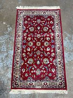 Top-down image of the entire diminutive rug showing red field with floral details and cream floral border with fringes at ends.
