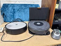 Two different iRobot Roomba robot vacuum cleaners on their charging docks, placed on a beige surface with a blue fabric background.