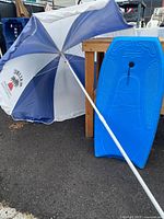 Blue Morey Boogie board resting against a wooden table with a vintage white and blue Malibu beach umbrella leaning beside it. Umbrella shows moderate fading on blue panels.