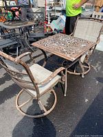 Photo of weathered metal patio table with cutout floral design, two swivel chairs with beige fabric seats, showing paint chipping and wear.