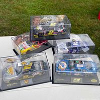 Five NASCAR diecast model cars on white table, each in clear display case with bases and featuring racing sponsor logos and numbers