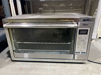Front view of Oster convection toaster oven showing glass door, stainless steel frame, digital display, and control buttons.