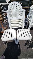 Photo showing a stack of 6 white plastic chairs with rounded backs and vertical slats along with 2 matching white square plastic tables with slatted tops.